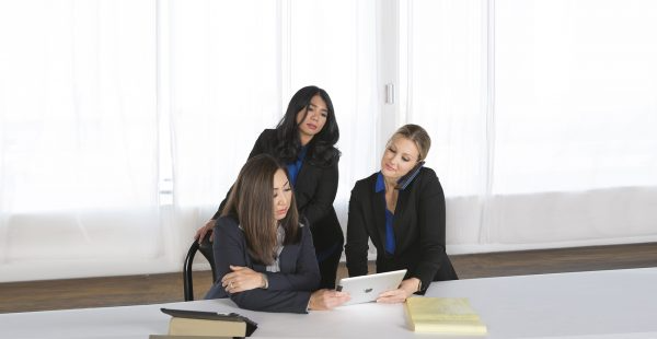 Three professional women collaborate in a bright office, gathering around a tablet while one speaks on a mobile phone.